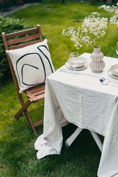 romantic dinner outside at summer, wooden chair with cushion, table serving with white linen tablecloth, empty plate and bowl, wine glass, fork, spoon and silver knife, vase with flowers on lawn