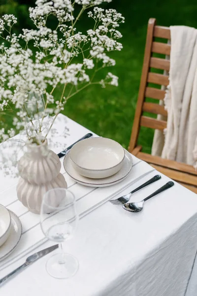 festive lunch outside at summer, table serving with white linen tablecloth, empty plate and bowl, wine glass, fork, spoon and silver knife, vase with flowers in green garden