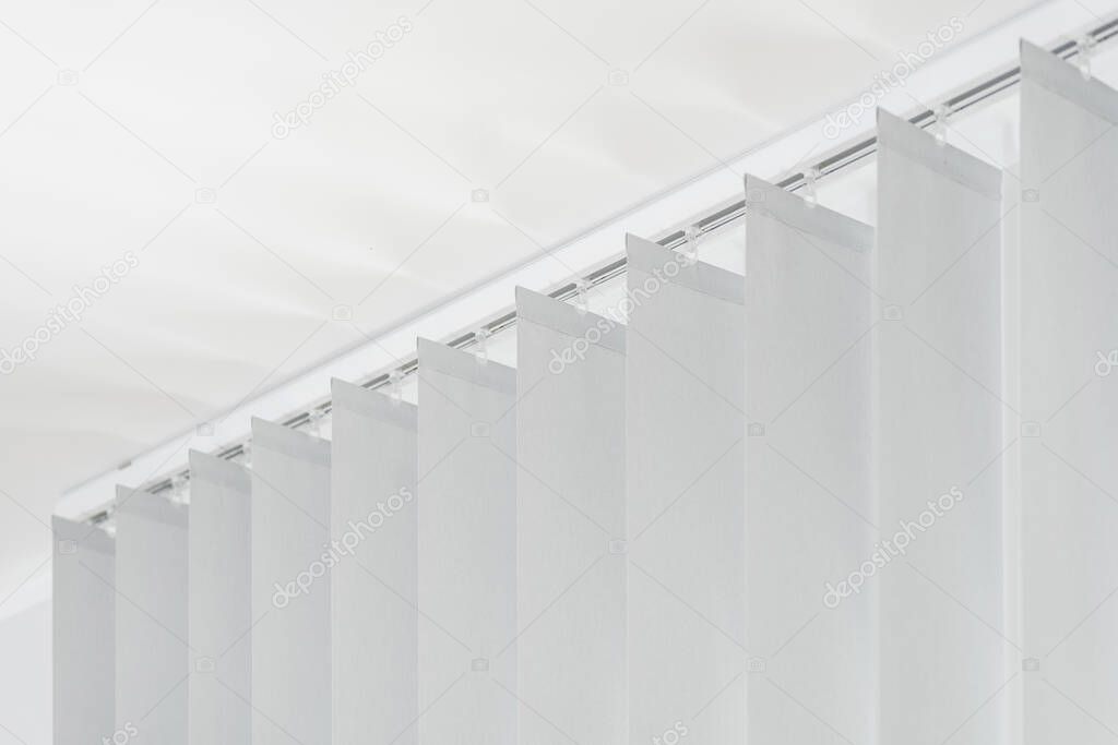 row of grey vertical blinds on ceiling cover sunlight from window in office. white fabric jalousie on headrail in room. household decor in apartment