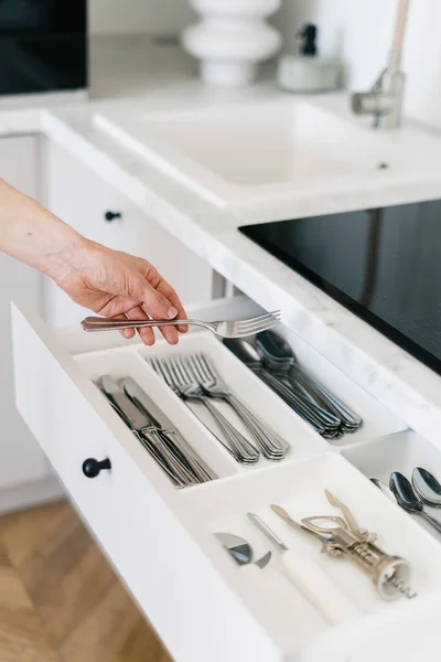 Cropped view of woman hold metal fork in hand, standing on kitchen near white open cutlery drawer. Housewife put clean and washed silverware in storage box, vertical shot