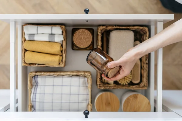 Top view of woman take recycled glass jar from open drawer in kitchen. Concept of organizing space at home. Storage box with towels, bamboo brush and cleaning sponge in wicker basket