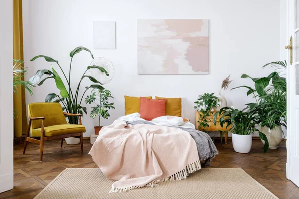 Wide open plan of light bedroom in apartment with retro interior design. Vintage style and colorful decor. Abstract art painting over bed with cushions, crumpled plaid, armchair and potted houseplants