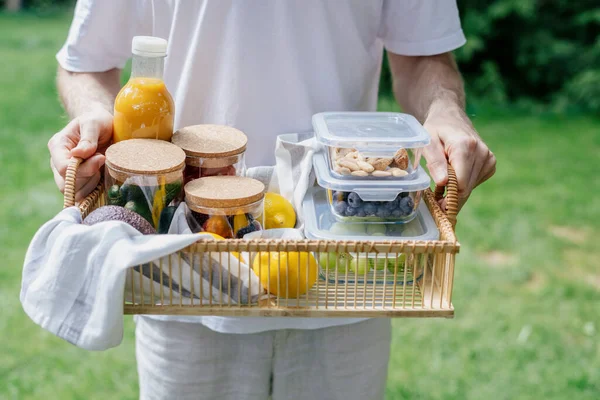 breakfast on backyard. garden picnic concept. rattan tray in male hands with berry and vegetables in boxes and glass containers, cropped shot