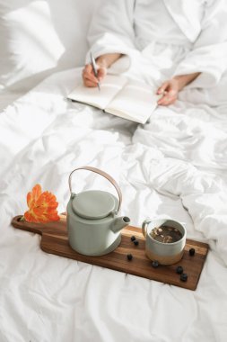 Sıcak bitki çayıyla birlikte kahvaltı tepsisinin dikey çekimi. Yatakta oturan, günlük tutan, sabahları evdeki rahat ve beyaz yatak odasında geçiren ya da otel odasında rahat eden bir kadın.