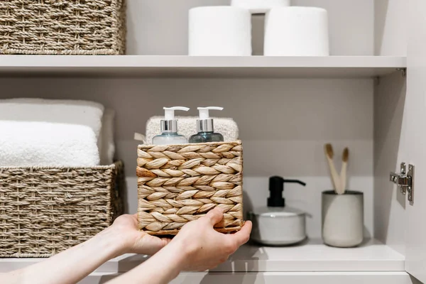 Organization of space in the bathroom cabinet. Cropped view of woman putting wicker box with bath sponge, shampoo, soap dispenser bottle and other cosmetics products in closet