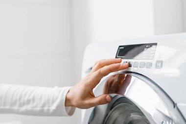 Kırpılmış kadın eli görüntüsü otomatik çamaşır makinesini açar ya da modern evdeki beyaz banyodaki kontrol panelinde düğmesi olan programı seçer