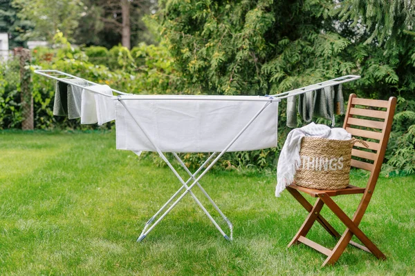 fresh terry towels and wet bed linen hanging on metal drying rack after laundry, wicker basket with clean clothes on wooden chair in backyard on green trees background