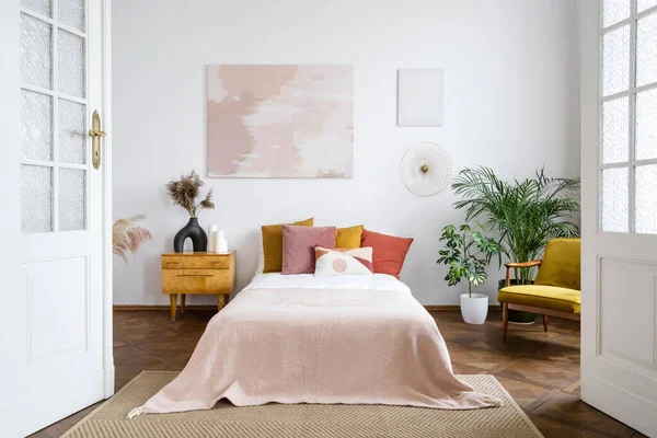 Vintage bedroom. Concept of interior design in style of the 70s. Wide open plan of cozy apartment with home decor, tidy bed, night table. Armchair and houseplants in room with abstract art painting