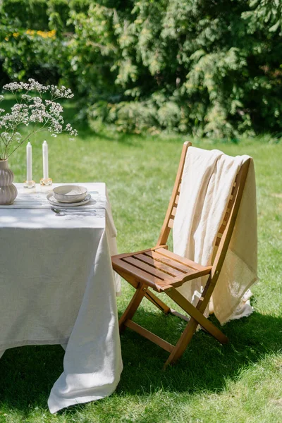 romantic breakfast in nature. wooden seat and table setting with white linen tablecloth, dinnerware, silverware, candles and flowers decor in vase in green park outdoors