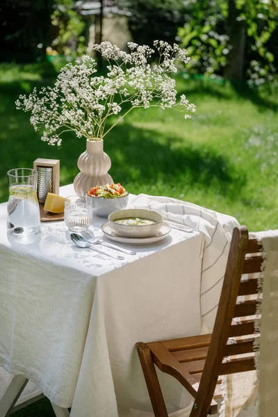 木製の椅子とテーブルは白いリネンのテーブルクロス 新鮮な野菜サラダ チーズ ボウルにスープ 冷たい水とレモングラスジュグと夏に屋外公園の花瓶の花を提供しています — ストック写真