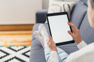 Kadın elinin kırpılmış görüntüleri taşınabilir dijital tablet cihazını tutuyor. Sahte ekranlı, oturma odasındaki kanepede okuyup iletişim kuruyor.