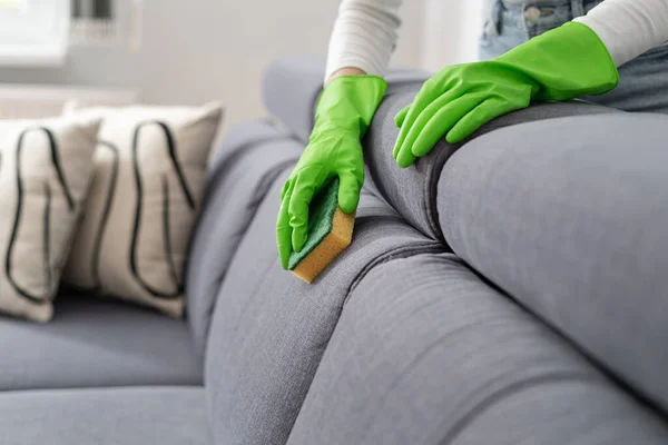 cropped shot of female hands in green rubber gloves wash couch upholstery with sponge in living room at modern apartment, housework concept