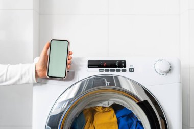 Beyaz banyoda kontrol paneli olan otomatik çamaşır makinesinin yanında fotokopi alanı olan kadın cep telefonu çekimleri, modern teknoloji konsepti.