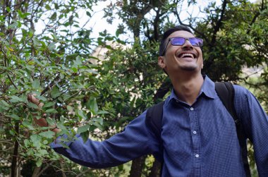 Adam parkta ağaçların ve çalıların arasında duruyor. Mutlu Hintli adam bir çalıyı tutarken dinleniyor.