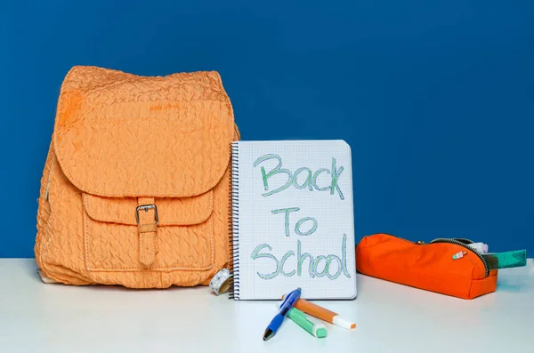 orange backpack with front pocket on blue background with markers and pen on white table. open notebook with 