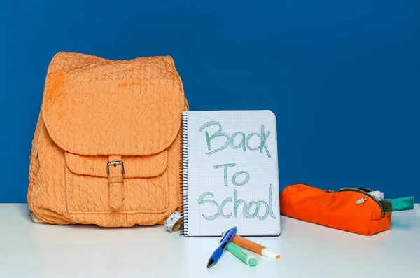 orange backpack with front pocket on blue background with markers and pen on white table. open notebook with \