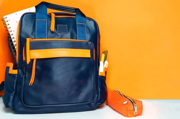 blue backpack with a front pocket on an orange background. an agenda protrudes and in the side pocket there are a few markers and a ruler. on the side there is a pencil case with an ice cream shaped pin. back to school concept with copy space.