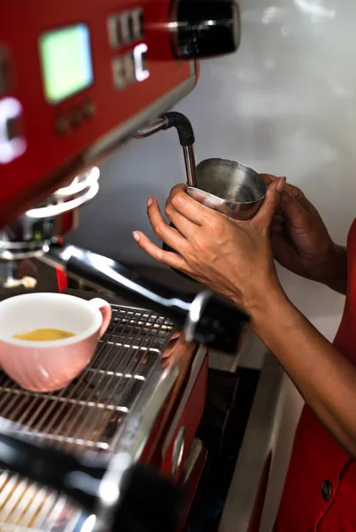 hands of a barista holding the metal jug where she is heating the milk for her customer\'s coffee. the machine and her uniform are red. there is a colorful cup with coffee. copy space. vertical photograph.