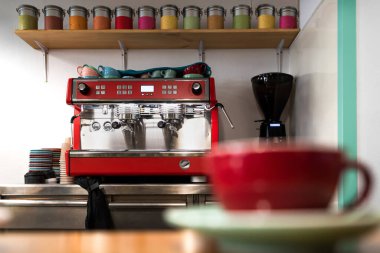 Ön planda odakları bozuk kırmızı bardağı olan küçük bir kahve dükkanındaki klasik profesyonel kahve makinesi. Tepesinde farklı renkte çay kutuları ve çeşitli renkli tabaklar var.