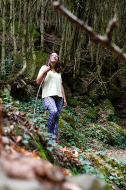 Genç yürüyüşçü ormanda duruyor. Bir sonbahar öğleden sonrasında ağaçların arasında poz veren bir kadın. Etrafındaki manzaraya bakarken destek için büyük bir dal tutuyor..