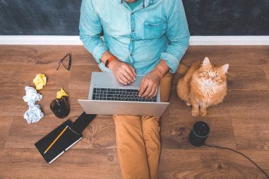 Yerde otururken evde çalışan bir adam. Ses kontrollü akıllı hoparlörün yardımıyla dizüstü bilgisayarda çalışıyorum. Zencefilli evcil kedili yaratıcı ofis. Amazon Alexa Echo Dot ile konuşuyor