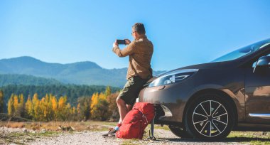 Adam arabayla seyahat ediyor ve cep telefonuyla fotoğraf ve video çekiyor. Yolculuk arasında mola ver. Araba sahipleri için cep telefonu uygulamaları. Kiralık arabayı mobil uygulamalarla kullanan bir adam. Dağlarla güzel doğa
