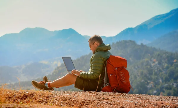 Hombre excursionista usando un ordenador portátil que trabaja en la cima de las montañas. Trabajo remoto con laptop. El hombre se sienta en la parte superior — Foto de Stock