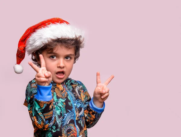 Mały, zabawny, latynoski chłopiec w świątecznym kapeluszu, uśmiechnięty ze szczęśliwą twarzą. Dziecko robi znak zwycięstwa. Dzieciak z przedszkola pokazuje numer dwa. Szczęśliwego Nowego Roku i Bożego Narodzenia — Zdjęcie stockowe