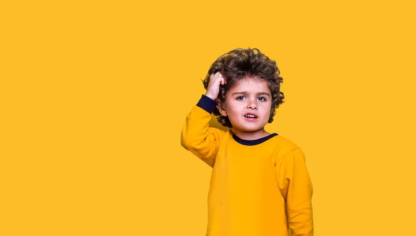 Smart funny curly kid scratches his head while thinking. Little wunderkind ready to go to school isolated on yellow background. Child looking up while thinking — Stock Photo, Image
