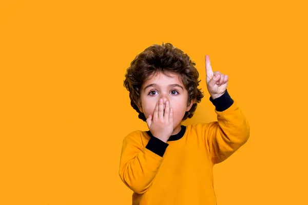 Little cute curly boy covering mouth with hand, shocked and amazed. Kid pointing up with finger isolated on yellow background. afraid for mistake. surprised expression — Stock Photo, Image