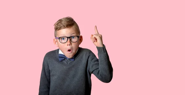 戴眼镜的滑稽傻小子指着手指向上看.令人惊讶的孩子有一个想法。用粉红的背景把食指竖起来.学校的书呆子 — 图库照片