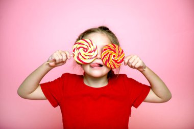 Gözlerinde şeker lolipopu olan komik çocuk, büyük şeker lolipopu yiyen mutlu küçük kız, şeker yiyen çocuk. Şekerli sürpriz çocuk. Pembe arka plan, stüdyo. Dilli komik çocuk.