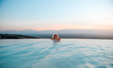 Outdoor luxury resort. Woman relaxing in nature swimming pool water. Sunset in the edge pool with beautiful view. Happy healthy female enjoying summer travel vacation. Relax, spa concept.