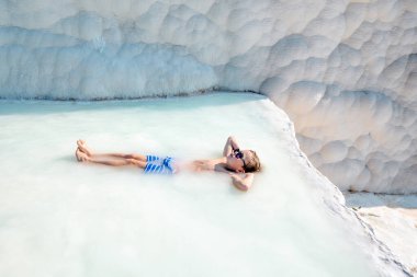 Little kid enjoy hot spring spa at Pamukkale while lying in hot spring water in Turkey. Pamukkale means cotton castle in Turkish. People have bathed in outdoor swiming pools. Child boy in sunglasses