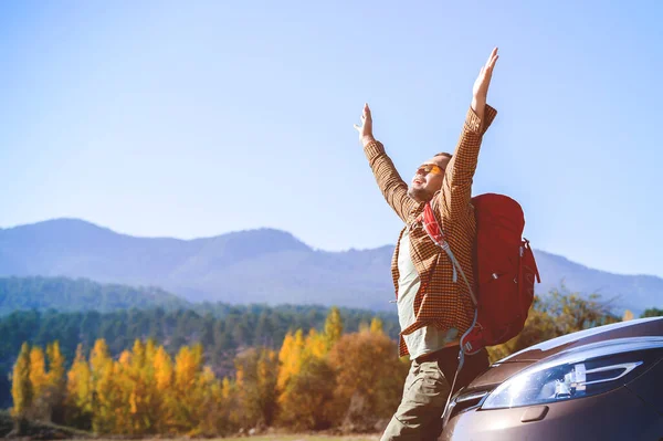 Man with arms raised up cheerful and happy sitting on car. Tourist with backpack enjoy mountain views.