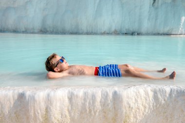 Yaz tatilinde güneş gözlüklü okul çocuğu. Rahatla ve kaplıcaya git. Çocuk açık havuzda dinleniyor. Beyaz kayalıklarda kaplıcalı lüks havuz