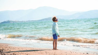 Deniz kıyısındaki çocuk. Güzel doğa. Güneşin altındaki sıcak kumda güneş gözlüklü yalnız çocuk. Çocuk dağların keyfini çıkar