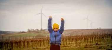 Yel değirmeninin önündeki okul çocuğunun portresi. Neşeli çocuk rüzgar türbinine bakıyor. Çevreyi keşfet. Enerji. Çocuk suratlı. Çocuk doğayı sever. Çocuk rüzgarla oynuyor.