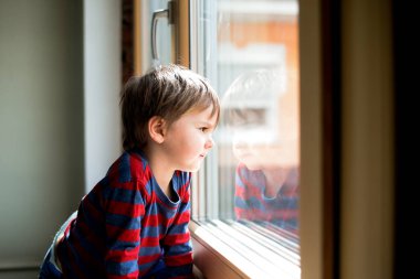 Kid boy sitting near window and looking outside. Serious child attentively looks out of the window clipart