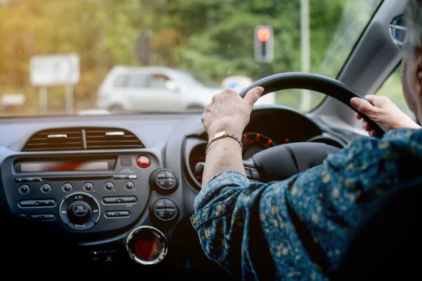 Elderly woman behind the steering wheel of a car. Cheerful senior woman driving a car. Old lady in glasses drive the car in glasses. Back view. Right wheel. England. UK.