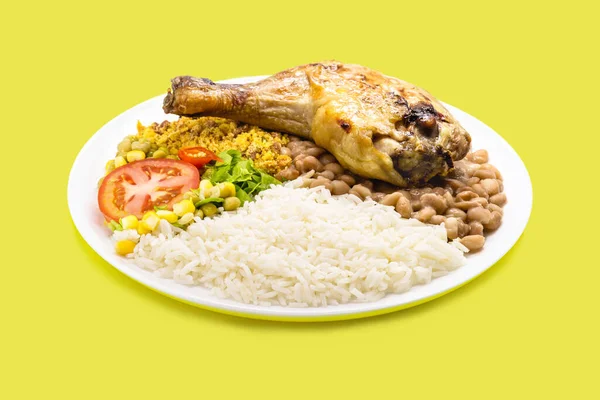 rice and beans dish, large roasted chicken thigh, chopped tomato salad with corn and peas, on isolated yellow background, traditional brazilian meal