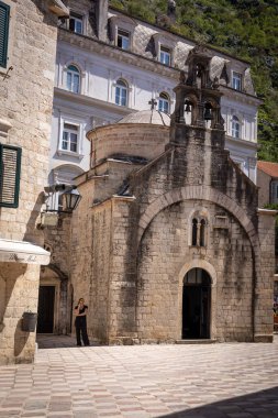 Kotor, Karadağ - 29 Nisan 2022: Kotor 'daki bir ortodoks Aziz Luke kilisesinin önü. Siyah giyinmiş bir kadın telefonda konuşuyor, duvarın önünde duruyor.. 