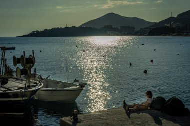 Budva, Karadağ - 28 Nisan 2022: Deniz kıyısındaki bir iskelede oturan çocuk. Suda yüzen küçük balıkçı tekneleri. Arka planda manzara dağları. 
