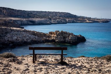 Kıbrıs Rum Kesimi, Cape Greco 'daki Blue Lagoon' da denize bakan boş ahşap bir bank. Küçük tekneler körfeze demir attı.. 
