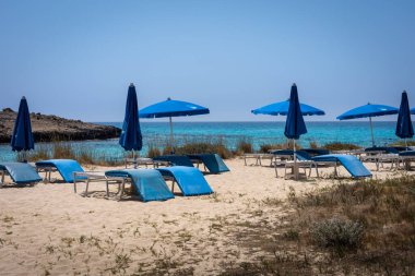 Ayia Napa, Cyprus - June 24, 2022: Blue umbrellas and empty sunbeds on the sandy beach with a sea view. 