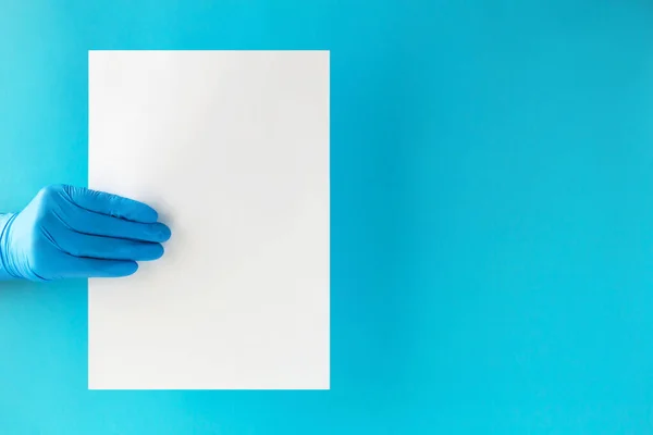 A doctors hand in a blue glove holds a sheet of white blank paper on a light blue background