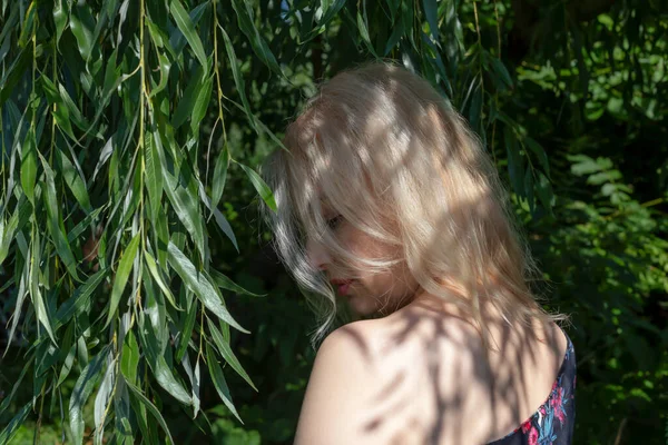 A blond girl stands in the foliage, half-turned, with her shoulder exposed. Her head is tilted, her eyes squinted. Beauty and thoughtfulness