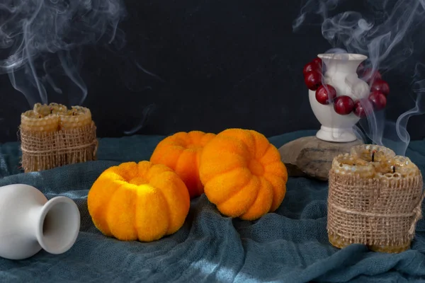 Halloween. There are pumpkins lying on a dark cloth, candles standing with smoke coming out of them, jugs and a red necklace. Black background with a copy space
