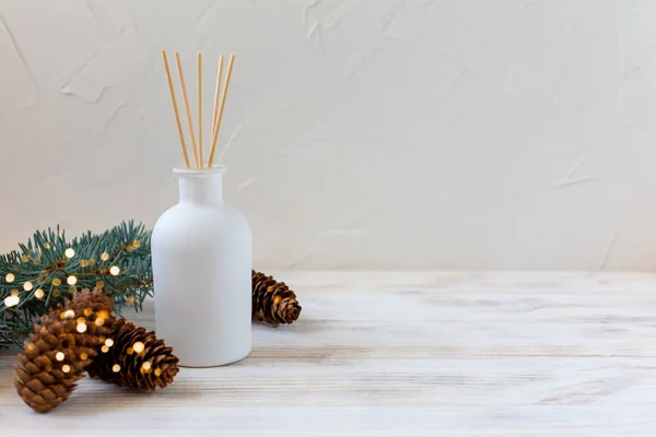 Perfumes for the home. Aromatic reed diffuser on a white background with a Christmas tree branch and cones in lights. The concept of peace, tranquility, relaxation and enjoyment