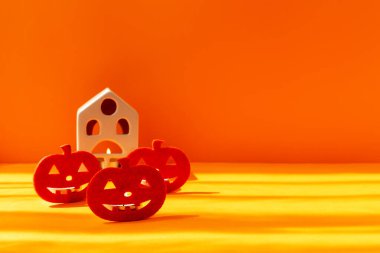 Cadılar Bayramı. Suni balkabakları güneş ışığında turuncu bir zemin üzerinde durur. Arkalarında yanan bir mum olan bir ev var.